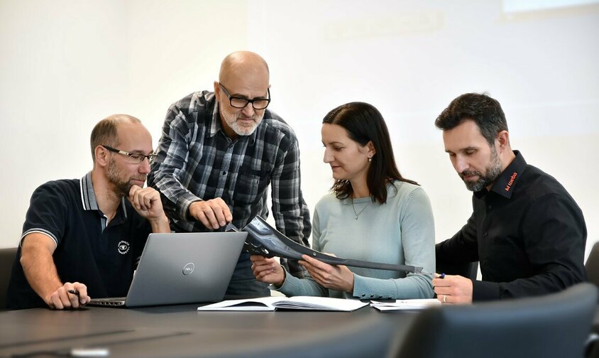 Tři zaměstnanci a jedna zaměstnankyně společnosti weba Werkzeugbau sedí na poradě o ocelové součásti. Na stole leží notebook a dokumenty.