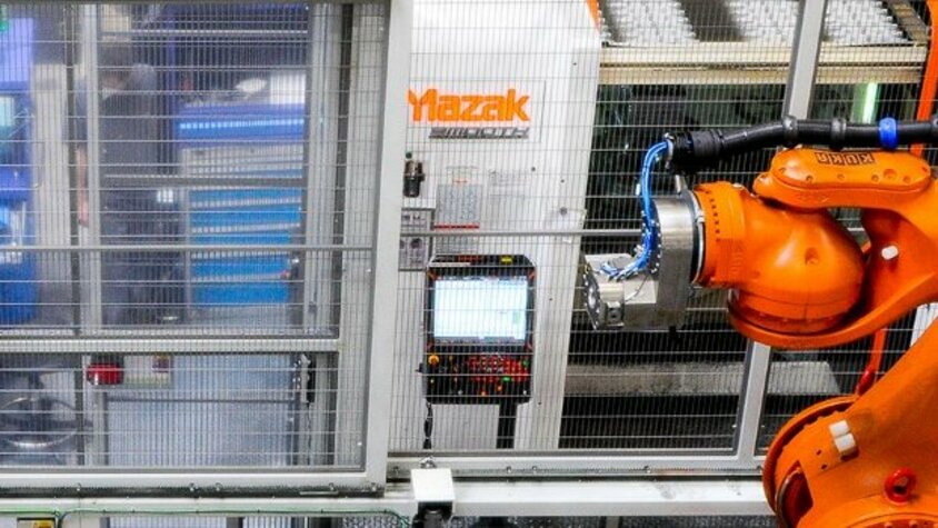 Zuverlässigkeit in der Lohnfertigung und mechanischen Bearbeitung Foto von zwei simultangesteuerten Mazak CNC-Bearbeitungszentren für die Lohnfertigung Fräsen, davor eine Transferstrecke mit einem Transferroboter. Die Aufnahme zeigt die hochpräzise Fertigungstechnik in einer modernen Produktionsumgebung, in der die Bearbeitungszentren komplexe Teile mit höchster Genauigkeit bearbeiten. Im Vordergrund ist die Transferstrecke zu sehen, die durch den Einsatz des Roboters einen effizienten Materialfluss zwischen den verschiedenen Stationen sicherstellt.