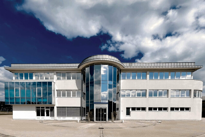 Exteriérový snímek administrativního křídla vývojového centra společnosti weba v Dietachu pod modrou oblohou s mraky.