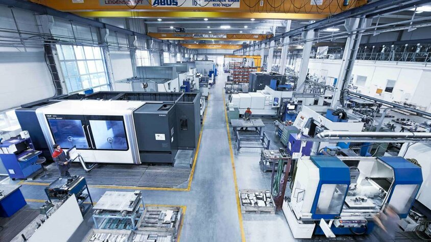 Moderne Produktionshalle für mechanische Bearbeitung Weitwinkelaufnahme einer modernen Produktionshalle für mechanische Bearbeitung. Mehrere CNC-Bearbeitungszentren und Maschinen sind in Betrieb, wobei Fachkräfte an verschiedenen Stationen arbeiten. Die Halle ist hell und gut organisiert, mit klaren Gängen für den Materialtransport. Im Hintergrund sind Kräne zur Handhabung schwerer Werkstücke sichtbar, was auf die hohe Kapazität der Fertigungseinrichtung hindeutet. Die Szene vermittelt Effizienz, Technologieeinsatz und ein professionelles Arbeitsumfeld in der mechanischen Bearbeitung.