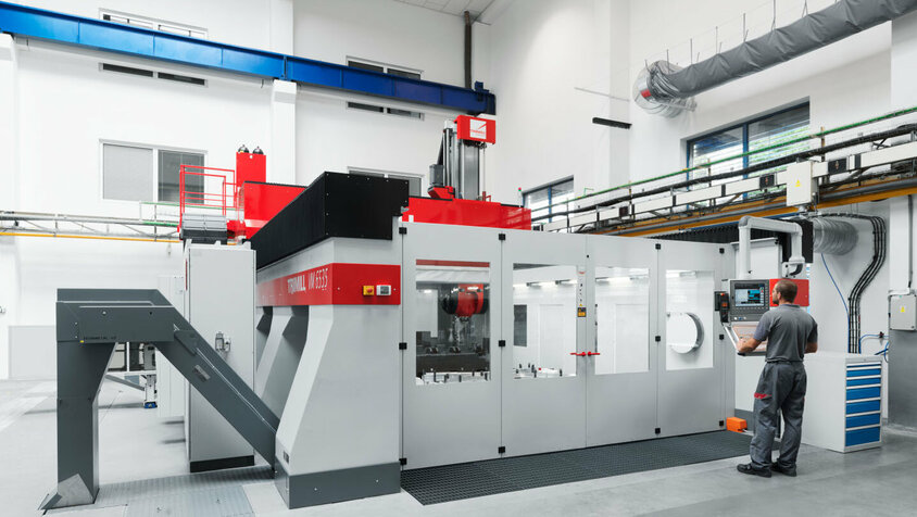 Hochmodernes CNC-Bearbeitungszentrum für mechanische Bearbeitung Ein Maschinenbediener arbeitet an einem modernen CNC-Bearbeitungszentrum für die mechanische Bearbeitung. Die Maschine ist in einer hellen und sauberen Produktionsumgebung installiert und verfügt über ein fortschrittliches Bedienfeld, das präzise Steuerung ermöglicht. Die robuste Bauweise der Maschine und ihre geschlossene Kabine deuten auf hochpräzise Bearbeitungsprozesse hin. Links neben der Maschine befindet sich eine Materialzufuhr, die den reibungslosen Ablauf des Bearbeitungsprozesses unterstützt.