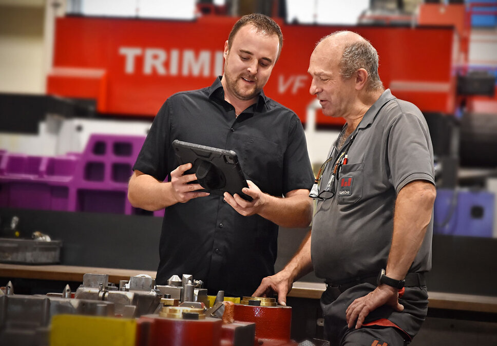 Ein Prozessmanager und ein Werkzeugbautechniker stehen in einer industriellen Umgebung und führen ein Gespräch über ein Werkzeug. Der Prozessmanager hält ein Tablet in der Hand und zeigt auf etwas auf dem Bildschirm, was anscheinend Gegenstand des Gesprächs ist. Im Hintergrund ist ein großes, rotes Bearbeitungszentrum von TRIMILL zu sehen, allerdings leicht verschwommen, um den Fokus auf die beiden Personen und ihre Interaktion zu legen. 