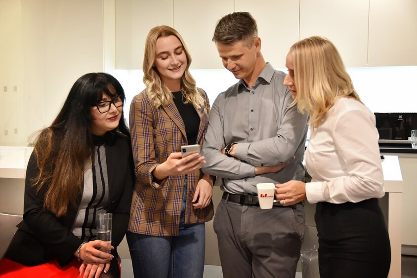 Tři zaměstnanci a jeden zaměstnanec společnosti Weba Olomouc stojí s úsměvem v kancelářské kuchyňce a společně se dívají na mobilní telefon. Dva lidé drží v ruce šálek kávy, což naznačuje uvolněnou, kolegiální atmosféru.