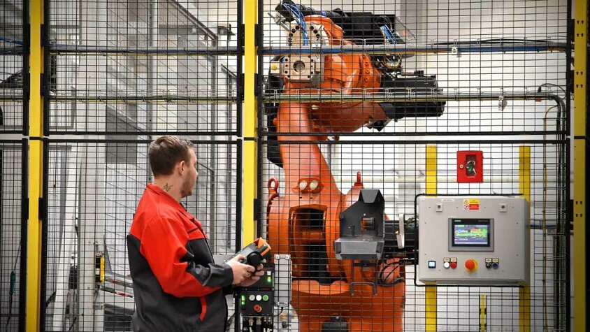 Bedienung eines Industrieroboters in der mechanischen Bearbeitung Ein Mitarbeiter in roter Arbeitskleidung bedient einen Industrieroboter in einer gesicherten Produktionszelle. Der Roboterarm ist hinter einem Sicherheitszaun sichtbar, und der Bediener hält ein Steuergerät in der Hand, um die Bewegungen des Roboters zu überwachen oder anzupassen. Neben dem Roboter befindet sich ein Bedienfeld mit einem digitalen Display, das zur Kontrolle und Überwachung der Roboterprozesse dient. Die Szene zeigt den Einsatz von Automatisierung und fortschrittlicher Robotik in der mechanischen Bearbeitung.