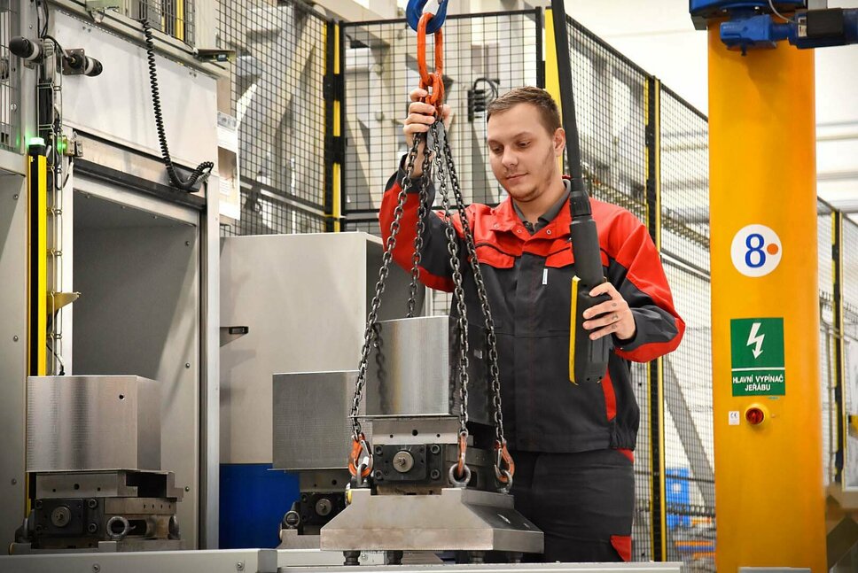 Obsluha stroje pracuje na přednastavovací stanici automatizovaného obráběcího centra pro smluvní výrobu. Pečlivě vkládá nástroje a součásti do seřizovacích stanic. Důraz je kladen na přesné umístění přesných nástrojů do určených pozic na složitém loži stroje. V pozadí je vidět průmyslové prostředí s vyspělou technologií.