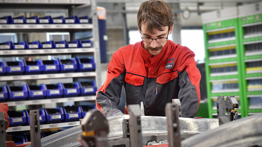 Werkzeug-Tryout - Langjährige Expertise bei weba Werkzeugbau Ein Werkzeugbautechniker baut konzentriert ein Presswerkzeug für anschließenden Werkzeug-Tryout und die Umformung von Strukturbauteilen zusammen; Im Hintergrund sind unscharf Regale mit Hilfs- und Betriebsstoffen