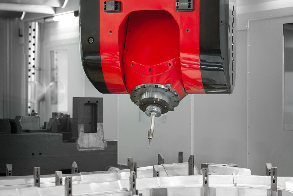 Detailní záběr CNC obráběcí hlavy při frézování v mechanickém zpracování. Červenočerná frézovací hlava je přesně umístěna nad upnutým obrobkem. Ostrý řezný hrot nástroje je připraven k vysoce přesnému obrábění, aby vyfrézoval do materiálu složité tvary. V pozadí je vidět interiér CNC stroje, který naznačuje kontrolované a profesionální výrobní prostředí.