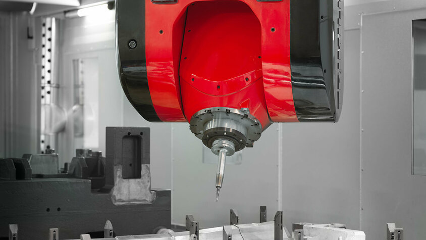 Präzisionsfräsen mit CNC-Technologie in der mechanischen Bearbeitung Nahaufnahme eines CNC-Bearbeitungskopfes während eines Fräsvorgangs in der mechanischen Bearbeitung. Der rot-schwarze Fräskopf ist präzise über einem eingespannten Werkstück positioniert. Die scharfe Schneidspitze des Werkzeugs ist bereit für die hochpräzise Bearbeitung, um komplexe Formen in das Material zu fräsen. Im Hintergrund ist der Innenraum der CNC-Maschine zu erkennen, was auf eine kontrollierte und professionelle Produktionsumgebung hindeutet.