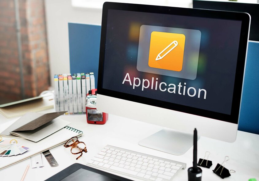  Pracovní stůl s obrazovkou Apple, klávesnicí a kancelářskými potřebami. Na obrazovce je vidět nápis „Application“, který symbolizuje volná pracovní místa v Olomouci ve společnosti weba Werkzeugbau.