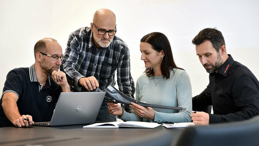 Jobs in Steyr - Karrieremöglichkeiten für Berufserfahrene bei weba Vier Personen sitzen an einem Besprechungstisch: Eine Person arbeitet an einem Laptop, eine andere betrachtet aufmerksam Unterlagen, während zwei weitere Personen sich angeregt über ein Stanzteil unterhalten. Die Szene zeigt eine konzentrierte und engagierte Arbeitsatmosphäre. Berufserfahrene und Profis finden bei weba ihren Traumjob in Steyr.