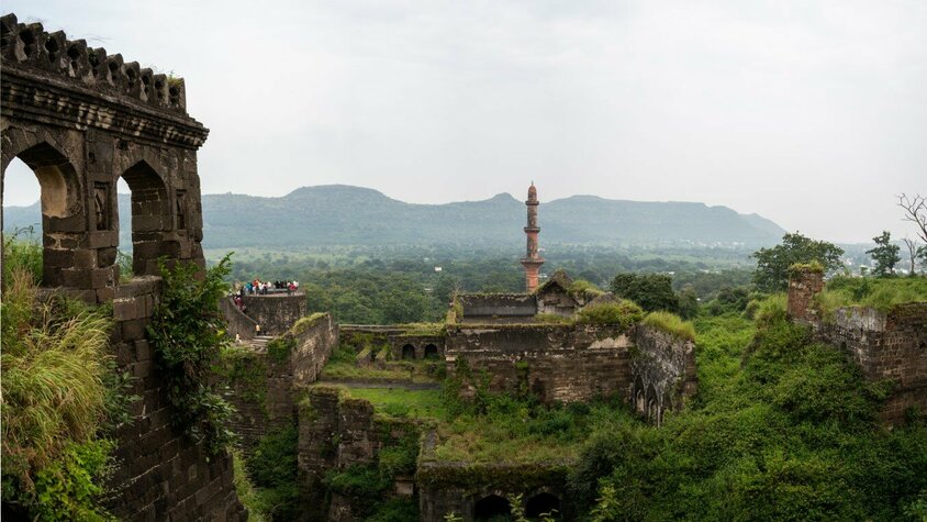 Historische Ruinen und grüne Landschaft: Eine Reise in vergangene Zeiten Eine historische Ruinenlandschaft mit üppiger Vegetation, einer alten Steinmauer im Vordergrund und einer hohen, markanten Säule im Hintergrund, eingebettet in eine weite grüne Landschaft.