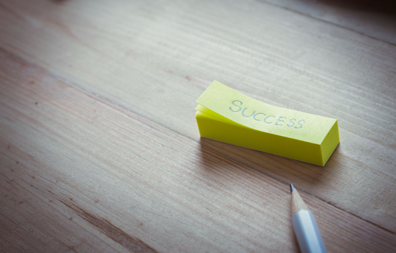 Gelber Notizzettel mit dem Wort „Success“ auf Holztisch, daneben ein Bleistift.